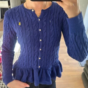 Blå kabelstickad kofta från Ralph Lauren - Säljer en blå kabelstickad kofta från Ralph Lauren med volang nedtill och vita knappar framtill. Klassisk gul logga broderad på bröstet. Rund halsringning och långärmad modell