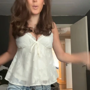 Vit blommig linnetopp med rosett - Säljer en söt vit linnetopp med blommigt mönster och liten rosett framtill. Toppen har v-ringning och tunna axelband, perfekt för varma dagar.