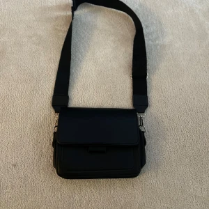 Svart axelväska med bred rem - Snygga svart axelväska med bred justerbar rem och stilren design. Väskan har ett lock framtill och flera fack för smart förvaring. Perfekt för att bära med sig det viktigaste i vardagen.