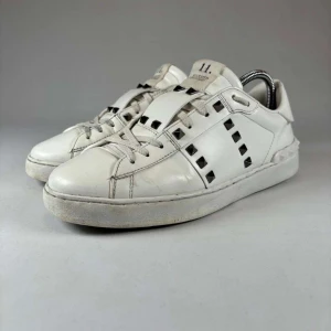 Vita Valentino sneakers med nitar - Säljer ett par vita Valentino sneakers med svarta fyrkantiga nitar längs sidorna och på plösen. Skorna har snörning och en klassisk rund tå. Tillverkade i skinn med en platt sula för en stilren look.