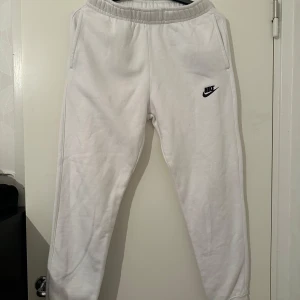 Vita mjukisbyxor från Nike - Säljer ett par vita mjukisbyxor från Nike med broderad logga på ena benet. Byxorna har resår i midjan och sidofickor, perfekta för en avslappnad stil.