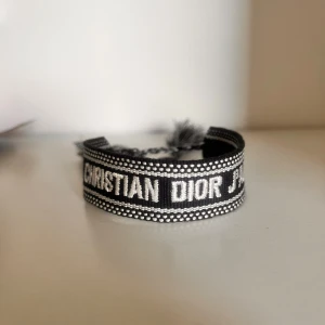Svart och vit armband från Christian Dior - Snyggt armband från Christian Dior i svart och vitt med tydlig logga broderad runt om. Armbandet har knytning med tofsar i ändarna och passar perfekt som en statement-accessoar.