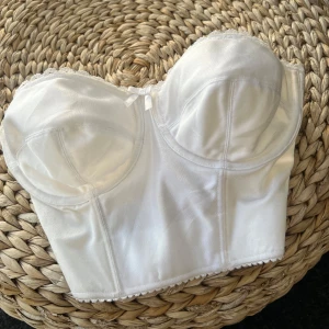 Vit korsett-topp från Vogue Bra - Säljer en vit korsett-topp från Vogue Bra i glansigt material. Toppen är axelbandslös med markerade kupor och bred resår i ryggen för extra stöd. Perfekt för att skapa en snygg siluett under kläder eller som statement-plagg.