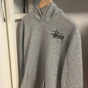 Grå hoodie från Stüssy - Säljer en grå hoodie från Stüssy med svart logga tryckt på både bröstet och ryggen. Köpt på plick för 200kr. Storleken står XS men den passar mer som en S/M. Fint skick och skön överlag.