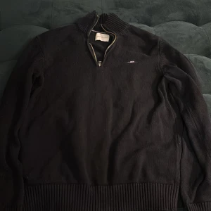 Svart stickad tröja med dragkedja från GANT - Säljer en svart stickad tröja från GANT med halv dragkedja vid halsen och broderad logga på bröstet. Tröjan har ribbade muddar och krage, perfekt för lager-på-lager. Klassisk och stilren design.