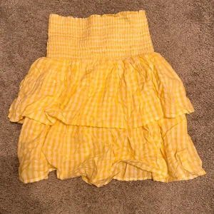 volangkjol - Superfin gul kjol med rutigt mönster och dubbla volanger. Kjolen har en smockad midja som ger en bekväm passform och är perfekt för varma dagar. Luftigt material och kort modell.