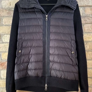 Moncler cardigan  - Säljer en svart pufferjacka från Moncler med stickade ärmar och quiltad front. Jackan har dragkedja, två fickor med tryckknappar och en extra ficka med dragkedja på ena ärmen. Perfekt för kyliga dagar.