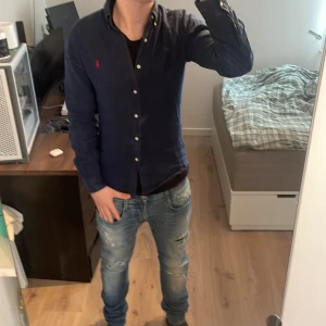 Mörkblå skjorta från Ralph Lauren - Säljer en mörkblå skjorta från Ralph Lauren med klassisk krage och knappar framtill. Skjortan har lång ärm och en liten röd broderad logga på bröstet. Perfekt för en stilren look.