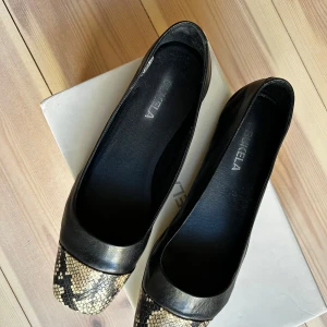 Svarta ballerinaskor med ormmönstrad tå från Bukela - Säljer ett par svarta ballerinaskor från Bukela med snygg ormmönstrad detalj på tån. Skorna har fyrkantig tå och är tillverkade i skinn med en platt sula. Perfekta för dig som vill ha något stilrent med en twist.