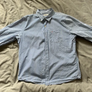 Blåvit randig skjorta från Hollister - Säljer en klassisk blåvit randig skjorta från Hollister med bröstficka och broderad logga. Skjortan har långa ärmar och knäppning framtill. Perfekt för en stilren look!