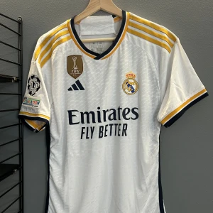 Real Madrid matchtröja Camavinga 12 - Snygg vit Real Madrid fotbollströja med guld- och marinblå detaljer, Adidas-logga och klubbmärke på bröstet. Nummer 12 och Camavinga på ryggen. FIFA-märke och UEFA Foundation-märke på ärmen. Klassisk V-ringning och korta ärmar. Helt ny med tagg en replika 