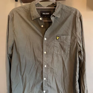 Olivgrön skjorta från Lyle & Scott - Säljer en olivgrön skjorta från Lyle & Scott i storlek M. Skjortan har klassisk krage, bröstficka med broderad gul logga och vita knappar framtill samt vid ärmslut. Perfekt för en stilren och avslappnad look.