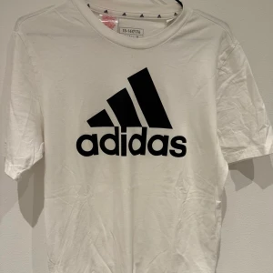 Vit t-shirt från Adidas med logga - Säljer en vit t-shirt från Adidas med stor svart logga och text på bröstet. Klassisk rund halsringning och korta ärmar. Perfekt för dig som gillar sportig stil och enkel design.