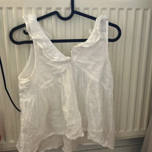 Vit volangprydd linnetopp från Gina Tricot - Säljer en söt vit linnetopp från Gina Tricot med volangdetaljer vid axlarna och knytning i ryggen. Toppen är ärmlös och har en luftig, lätt struktur – perfekt för varma dagar.