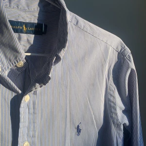 Blårandig skjorta från Ralph Lauren - Klassisk blårandig skjorta från Ralph Lauren med button-down krage och broderad logga på bröstet. Skjortan har långa ärmar och vita knappar framtill. 10/10 skick, perfekt för både daglig användning och fest.