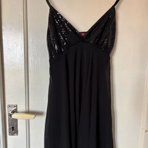 Svart paljettklänning från John Zack - Säljer en svart festklänning från John Zack med smala axelband och paljetter upptill. Klänningen har v-ringning och slutar ungefär mitt på låret.