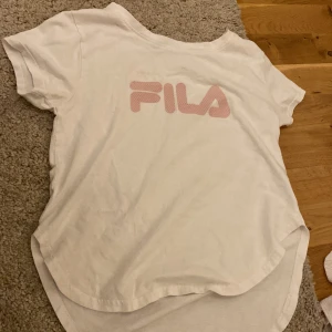 Vit t-shirt från FILA med rosa logga - Vit t-shirt från FILA med stor rosa logga på bröstet. Klassisk rund hals och korta ärmar. Perfekt för en sportig och avslappnad stil. Tillverkad i mjuk bomull för extra komfort.