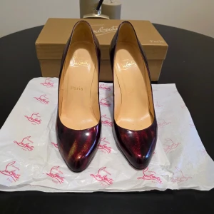 Vinröda pumps från Christian Louboutin - Säljer ett par klassiska vinröda pumps från Christian Louboutin med den ikoniska röda sulan. Skorna har spetsig tå och 100 mm hög klack, tillverkade i skinn med en glansig finish. Rekommenderar att du är en storlek 37/38 i vanliga fall då de är väldigt små i storleken