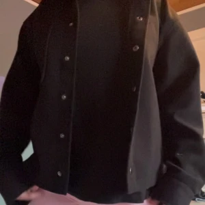 Svart kappa med knappar - Säljer en svart vår kappa/jacka med klassisk krage och tryckknappar framtill. Jackan har en bröstficka och är i ett mjukt material som känns bekvämt. Perfekt att slänga över en t-shirt för en enkel och stilren look. Endast använd 1 gång som ny. storlek 158/164 men passar mig som kan ha xs-m💕