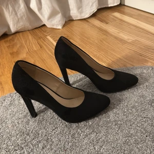 Svarta klackar från Graceland - Klassiska svarta klackar från Graceland med hög klack och spetsig tå. Skorna har en elegant och tidlös design som passar till många olika outfits. Utsidan är i ett mjukt material och insidan är ljusbeige.