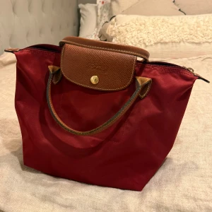 Vinröd handväska från Longchamp - Vinröd handväska från Longchamp med bruna skinnhandtag och lock. Väskan har dragkedja och knappstängning i guldton. Har en liten blekningsfläck men den syns knappt. ❤️