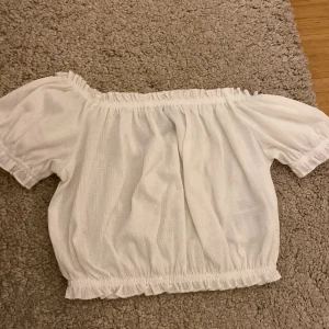 Vit offshoulder croppad topp från H&M Divided - Säljer en vit croppad offshoulder topp från H&M Divided i storlek XS. Toppen har resår vid axlar och midja för en bekväm passform och korta ärmar. Perfekt till sommaren!