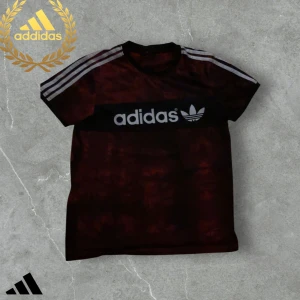 Röd Adidas t-shirt med vita ränder - Snygg röd t-shirt från Adidas med svart panel och stor vit logga framtill. Klassiska tre vita ränder på axlarna och korta ärmar. Perfekt för en sportig look.