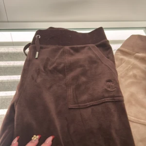 Bruna mjukisbyxor från Juicy Couture - Mysiga bruna mjukisbyxor från Juicy Couture med resår och snörning i midjan. Byxorna har fickor fram och bak, samt en broderad Juicy-logga på ena fickan. Perfekta för en avslappnad stil.