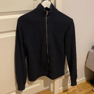 Svart stickad tröja med dragkedja från Dobber - Säljer en svart stickad tröja från Dobber i storlek small. Tröjan har hög krage och hel dragkedja framtill. Ribbstickad struktur och långa ärmar, perfekt för kyligare dagar