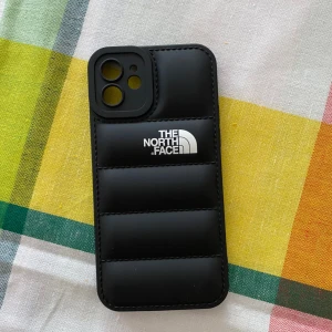 Svart mobilskal från The North Face - Säljer ett svart mobilskal från The North Face med vadderad design och logga på baksidan. Skalet har en quiltad look och passar perfekt för dig som vill ha en snygg och sportig accessoar till mobilen. Passar IPhone 12. Ej äkta 