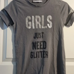 Grå t-shirt till flicka med glittertext från Red & Blue - Grå t-shirt från Red & Blue med texten 'GIRLS JUST NEED GLITTER'. T-shirten har korta ärmar med en dekorativ detalj och rund halsringning. 