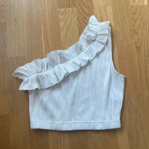 Vit one shoulder topp med volang - Säljer en vit one shoulder cropped topp med volangdetalj längs axeln. Toppen har en ribbad struktur och är ärmlös, perfekt för varma dagar eller en snygg look. Aldrig använd!
