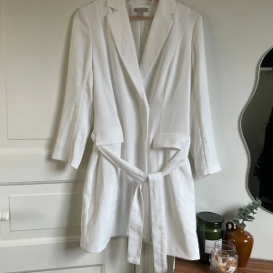 Vit kavajklänning med knytband  - Säljer en stilren vit kavajklänning från H&M med knytband i midjan och klassisk krage. Klänningen har långa ärmar och två fickdetaljer framtill. Perfekt för en chic look till studenten.🤍Lös tråd, se bild 5. 