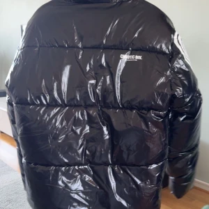 Svart pufferjacka från Chaotic Div. - Snygg svart pufferjacka från Chaotic Div. med glansig yta och dragkedja framtill. Jackan har långa ärmar och en cool, oversized passform. Perfekt för att hålla värmen med stil.