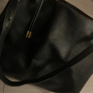 Svart axelväska med dragkedja - Snygg svart axelväska i fejk skinn med dragkedja och gulddetaljer. Perfekt för att bära med sig det nödvändigaste. Enkel och stilren design som passar till många tillfällen.