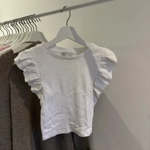 Vit topp från Zara - Säljer en söt vit ribbad topp från Zara med puffiga korta ärmar. Perfekt för en stilren look. Toppen är croppad och har en rund halsringning.