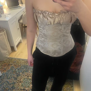 Beige korsett med spetsdetaljer - Elegant beige korsett med vackra spetsdetaljer och snörning i ryggen. Den har en rynkad överdel som ger en romantisk känsla. Perfekt för att ge en stilfull touch till din outfit.
