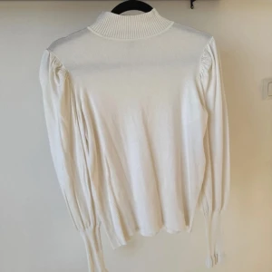 Vit stickad polotröja - Säljer en stilren vit polotröja med ribbad design och långa ärmar. Tröjan har en hög krage och puffiga ärmar som ger en elegant touch. Perfekt för kyligare dagar. Tröjan är storlek 14 - skulle säga att den motsvarar en L. 