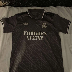 Real Madrid 2024/2025 fotbollströja  - Säljer en grå fotbollströja från Adidas med korta ärmar och tryck på både fram- och baksida. Tröjan har en klassisk krage och är dekorerad med klubbens emblem och sponsorlogga. Perfekt för fotbollsfans!