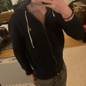 Mörkblå  hoodie från Ralph Lauren - Säljer en stilren blå hoodie från Ralph Lauren.           Enda defekten är på dragkedjan då själva kroken har lossnat men den fungerar som normalt och annars inget fel på den.