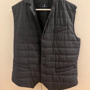 Grå quiltad väst från Hansen & Jacob - Snygg grå quiltad väst från Hansen & Jacob med knappar framtill. Perfekt för lager-på-lager-stil. Västen har en klassisk krage och två fickor framtill för extra stil och funktionalitet.