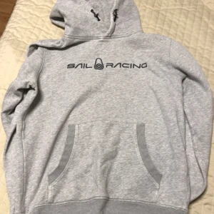 Grå hoodie från Sail Racing - Säljer en grå hoodie från Sail Racing med tryck på bröstet. Den har en klassisk känguruficka och justerbar huva med dragsko. Perfekt för en avslappnad stil.