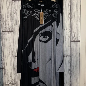 Svart och grå klänning med ansiktsmotiv från Tendency - Unik långklänning från Tendency med ett stort ansiktsmotiv i svart och grått. Klänningen har spetsdetaljer vid halsen och långa ärmar. Perfekt för dig som vill sticka ut med en konstnärlig touch.