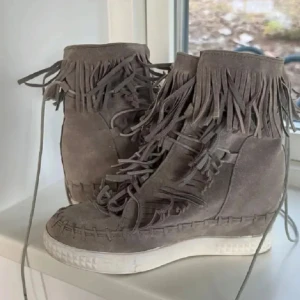 Fransskor - Snygga grå boots i mocka med fransdetaljer och snörning. De har en vit sula och en kilklack som ger extra höjd. Perfekta för en trendig look.