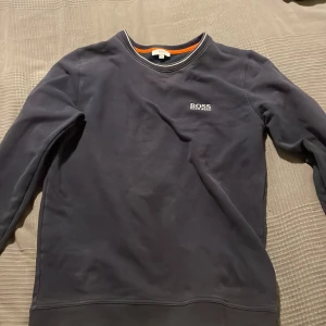 Mörkblå tröja från Hugo Boss - Säljer en stilren mörkblå tröja från Hugo Boss med rund halsringning och subtil logga på bröstet. Tröjan har långa ärmar och en kontrasterande orange insida vid halsen. Perfekt för en avslappnad och snygg look.