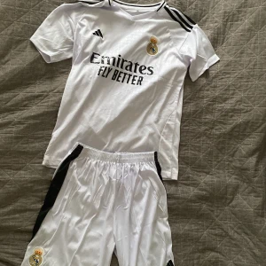 Vit Real Madrid fotbollströja - Säljer en vit Real Madrid fotbollströja från Adidas med korta ärmar och svarta detaljer. Tröjan har klubbens emblem på bröstet och sponsorlogga framtill. Perfekt för alla fotbollsfans som vill visa sitt stöd för laget!