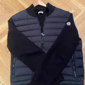 Moncler Cardigan - Snygg svart jacka från Moncler med dragkedja och ribbad krage. Jackan har långa ärmar och två fickor med knappar framtill. Perfekt för kyligare dagar. Sparsamt använd skick 9/10. Taggar ingår. Storlek L men passar M. Säljs då den inte längre passar