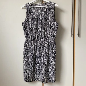 Svartvit mönstrad klänning från Miss Milla - Svartvit klänning från Miss Milla med ett geometriskt mönster. Klänningen är ärmlös och har rymliga fickor med dragkedjor på sidorna. Längd 93 cm, maxvidd runt midjan 110 cm. Tunn, sval polyester. 