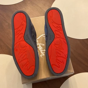 Grå sneakers från Christian Louboutin - Snygga grå sneakers i mocka från Christian Louboutin med den ikoniska röda sulan. Skorna har snörning och en stilren design som passar perfekt för en trendig look.