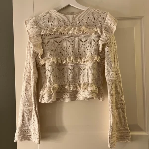 Vit spetsblus med volanger - Charmig vit spetsblus med vackra volangdetaljer. Blusen har ett intrikat spetsmönster och långa ärmar, perfekt för en romantisk look. Passar utmärkt till både jeans och kjol för en stilfull outfit.